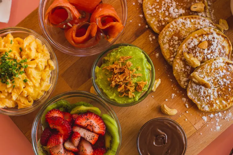 Tela com variados alimentos, incluindo salada de morangos, abacate picado com cobertura de bacon, ovos mexidos, pão doce...