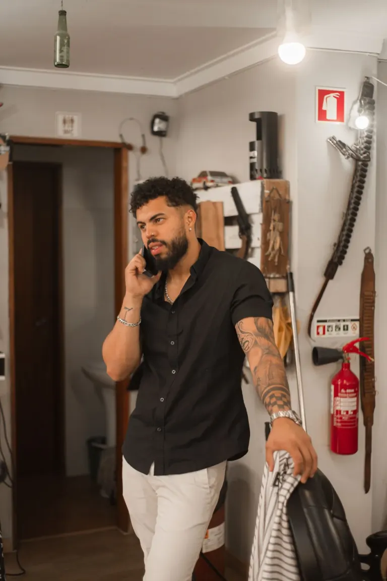 Homem com barba e tatuagens, usando camisa preta e calças claras, fica em um ambiente com ferramentas de cozinha na...