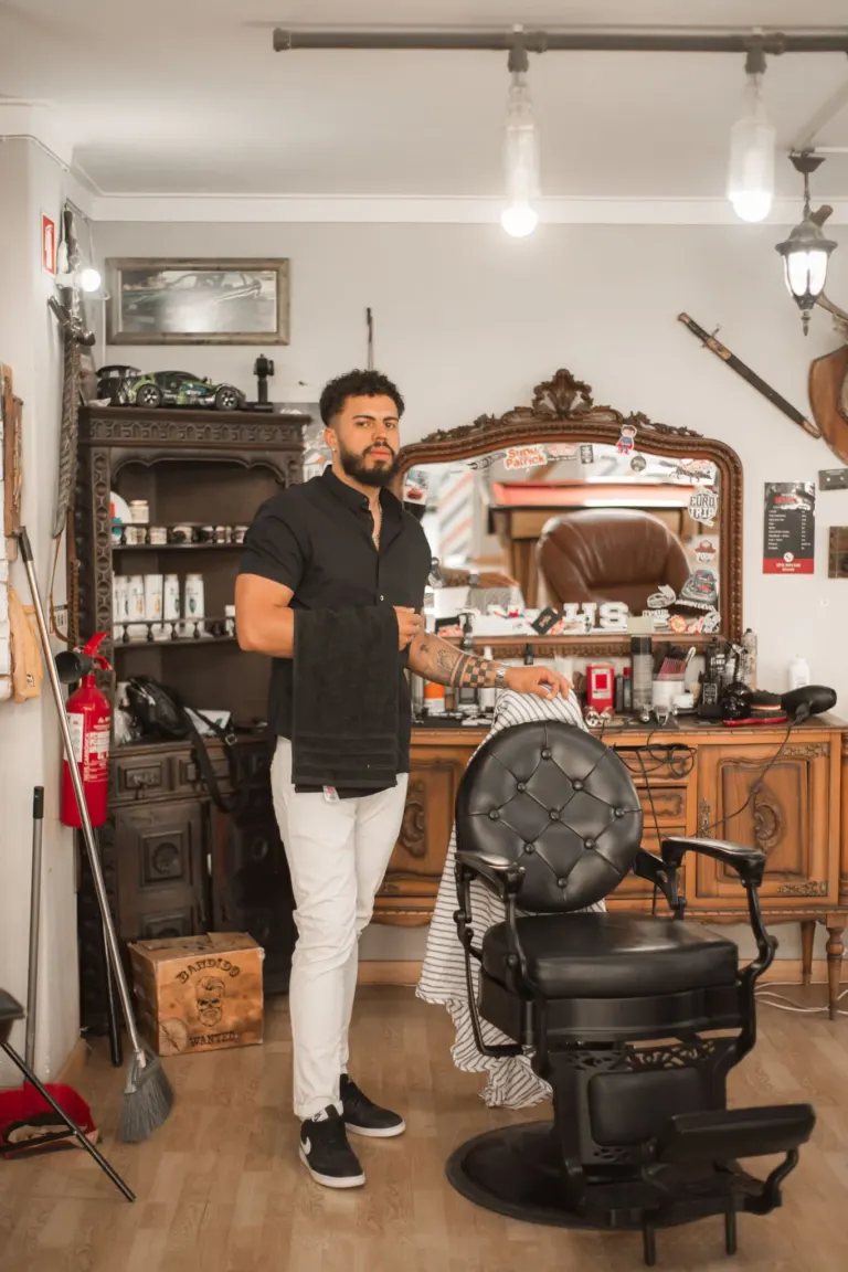 Homem de cabelo cacheado, barba, usando camiseta preta, calça branca e tênis preto, segurando uma toalha, dentro de uma...