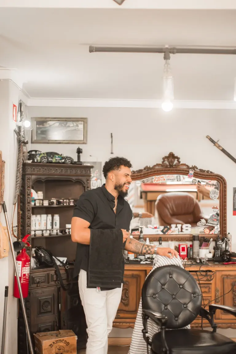 Homem sorrindo em barbeiro, segurando uma toalha preta, ao lado de uma cadeira de barbeiro e espelho decorado com acessórios