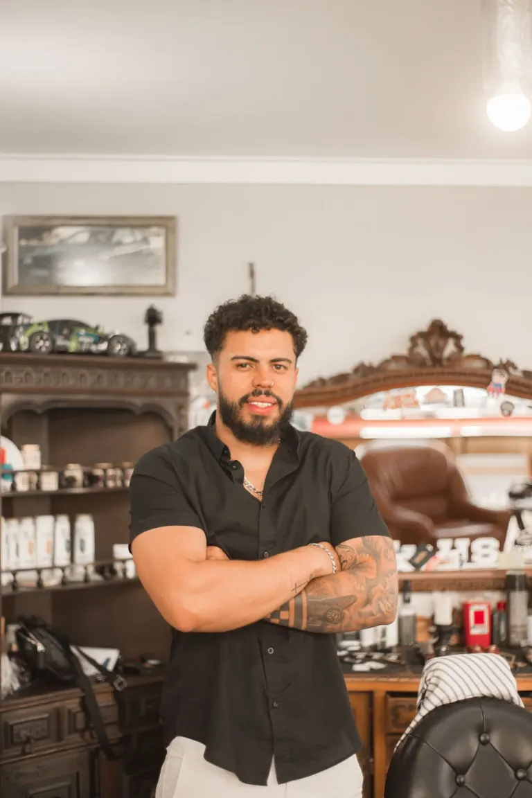 Homem de cabelo cacheado, barba e tatuagens no braço, vestindo camisa preta, posando com os braços cruzados em barbearia...