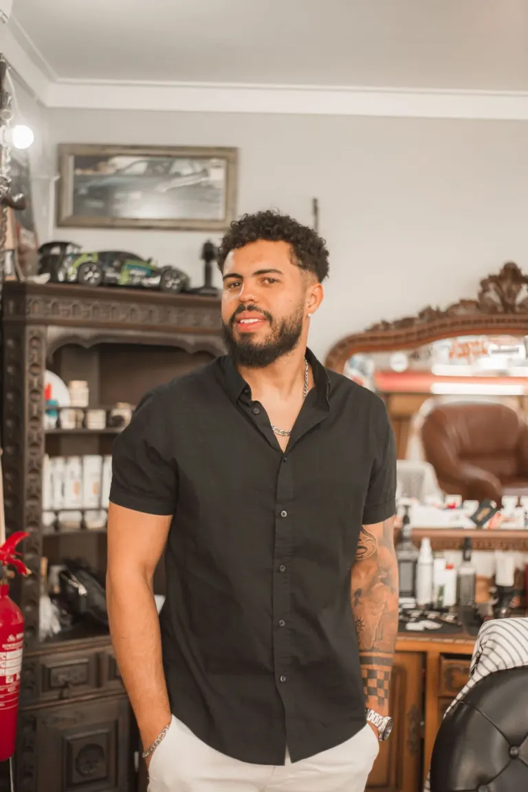 Homem com barba e cabelos cacheados, vestindo camiseta preta e calça branca, posando em barbearia com móveis antigos e...