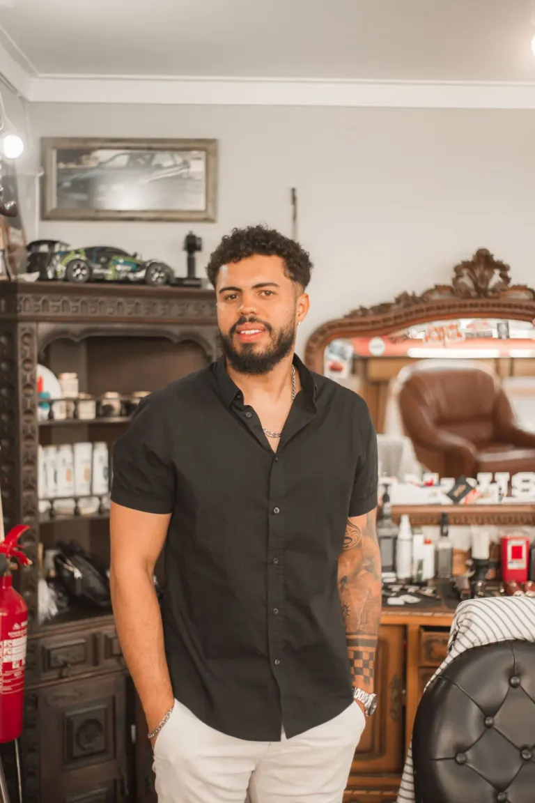 Homem de cabelo cacheado, barba e tatuagens, usando uma camiseta preta e calça bege, posando em um ambiente com móveis...