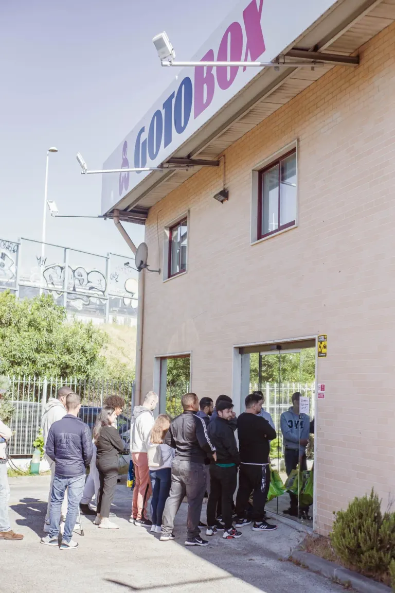 Fila de pessoas esperando para entrar em uma loja com uma placa azul e roxa que diz 'LOJAS'.