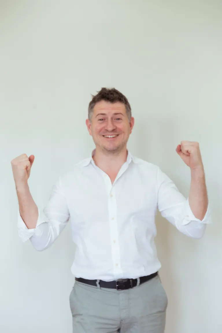 Homem sorridente vestindo camisa branca e calça cinza, levantando os braços em sinal de vitória ou conquista.
