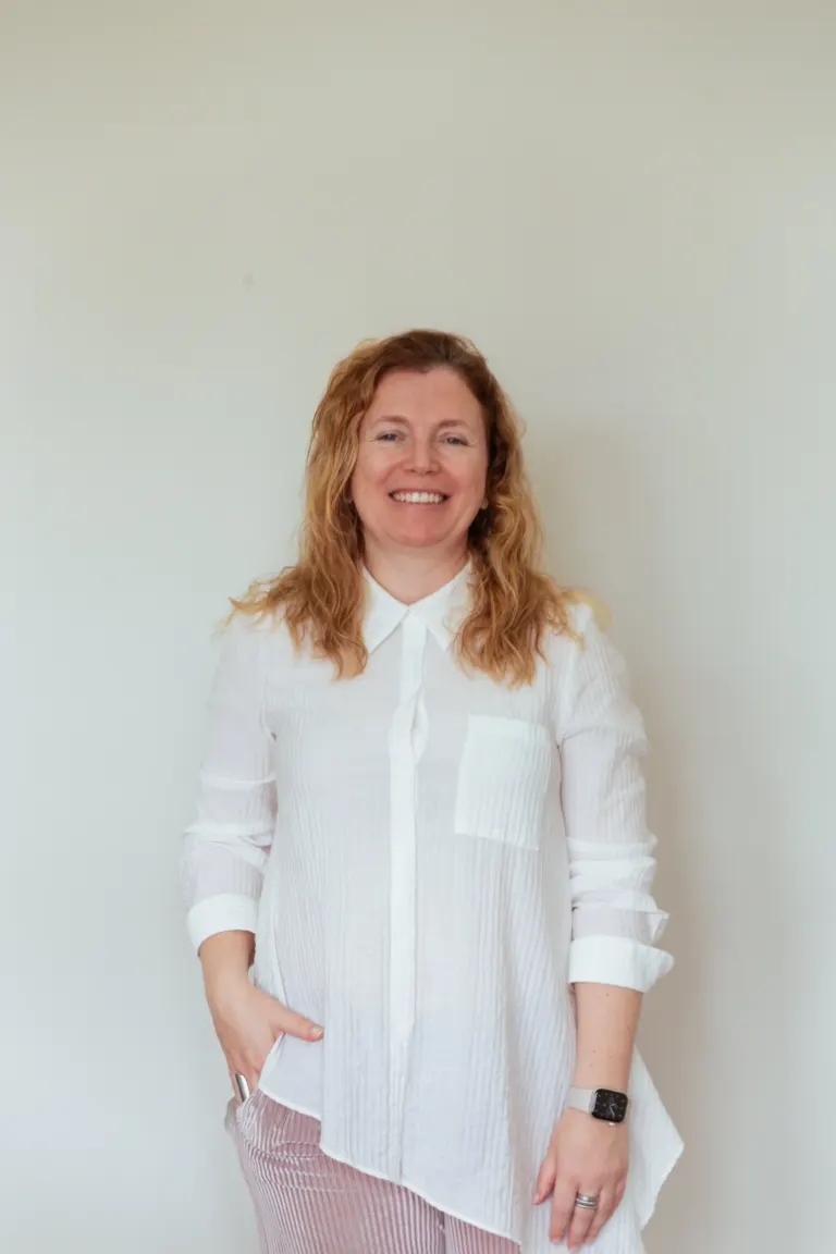 Mulher sorrindo com cabelo castanho ondulado, usando camisa branca de manga comprida e uma roupa listrada, de pé contra...