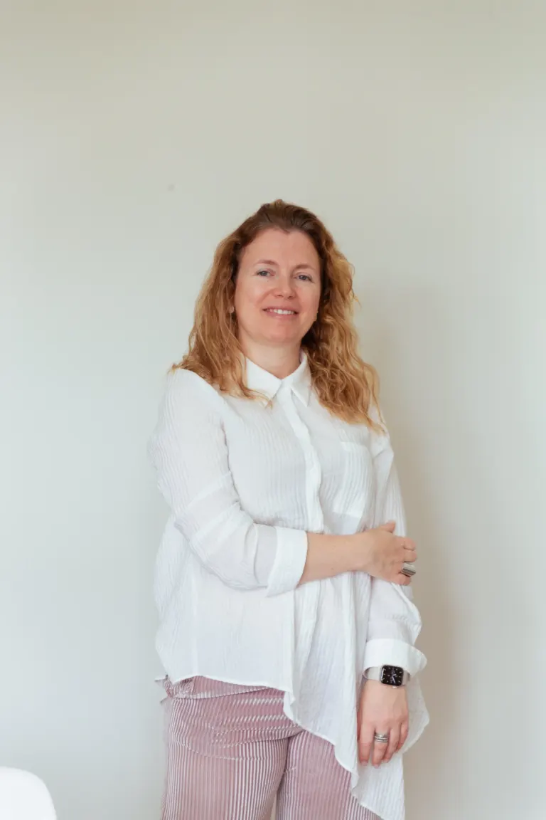 Mulher com cabelo castanho claro, usando camisa branca e calças risca de giz, sorrindo, em ambiente interno com parede...