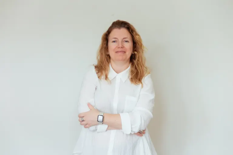 Mulher com cabelo castanho claro, usando blusa branca com mangas 3/4 e um smartwatch na esquerda, cruzando os braços,...