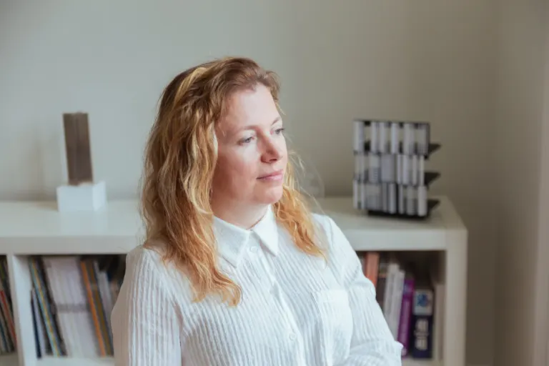 Mulher com cabelo ruivo sentado em ambiente de escritório ou sala com armários e livros ao fundo.