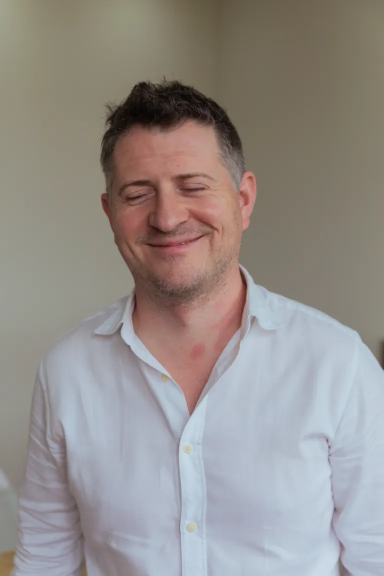 Homem sorridente com cabelo castanho, penteado para trás, usando camisa branca com botões, fechada na parte inferior, em...