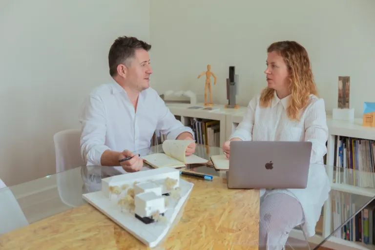 Homens em reunião de trabalho, com livros, laptop e modelo de projeto no mesa.