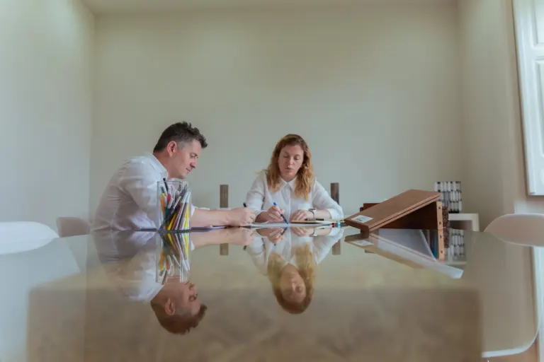 Homens e mulher sentados à mesa fazendo anotações, com uma jarra de lápis na mesa, em uma sala de reuniões