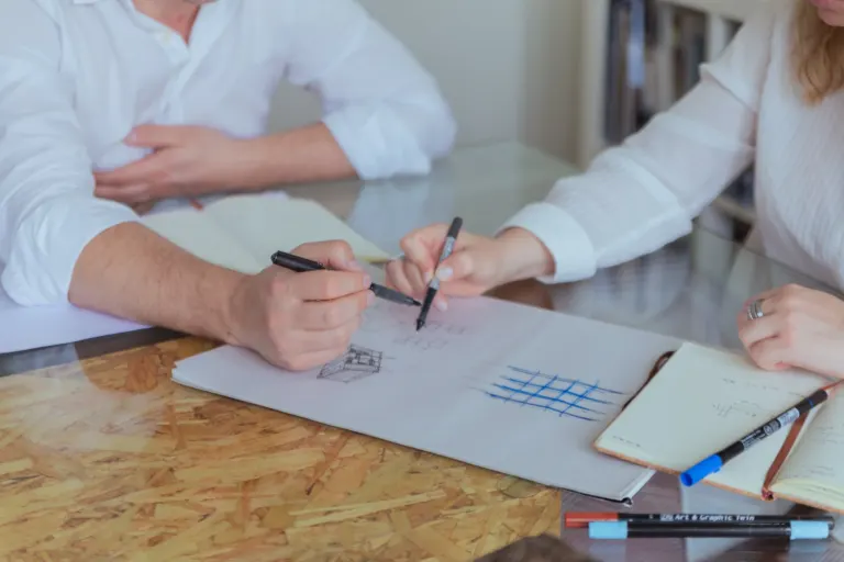Pessoa desenhando e fazendo anotações em uma mesa de madeira, com cadernos e canetas ao redor.
