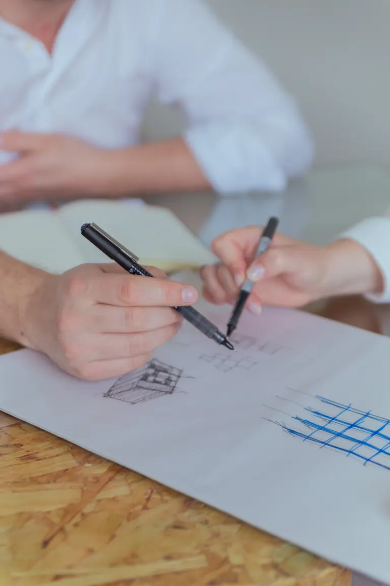 Duas pessoas analisando desenhos técnicos e de arquitetura, com uma delas segurando duas canetas pretas.