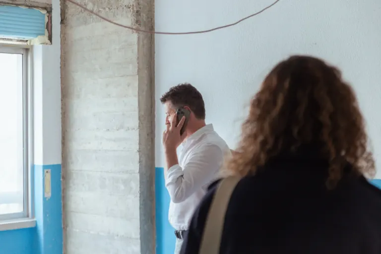 Homem com roupa social falando ao telefone em um ambiente que parece uma obra ou reforma, com parede parcialmente pintada...