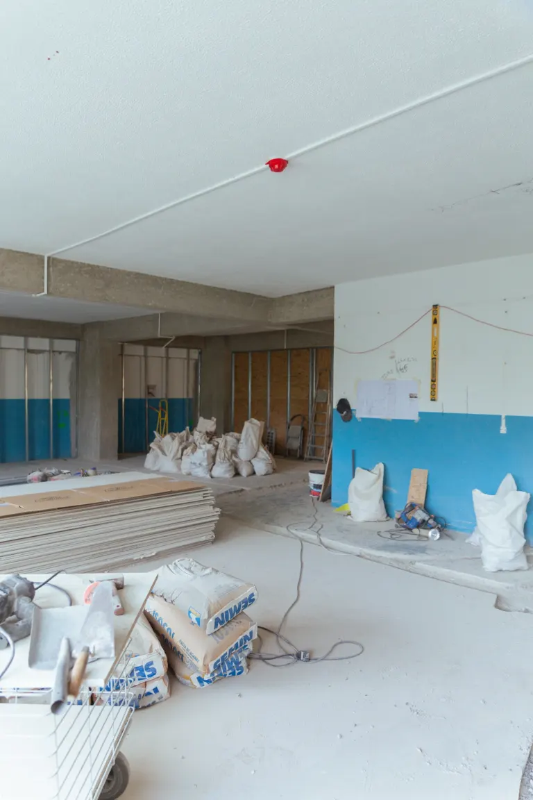 Interior de um local em construção com materiais, ferramentas e sacos de cimento, paredes em fase de acabamento e teto...