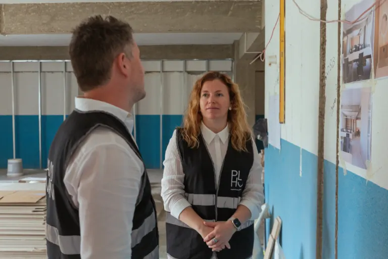 Duas pessoas discutindo em um local de construção, uma mulher com cabelo cacheado ruivo usando colete com logo "PLH" e um...