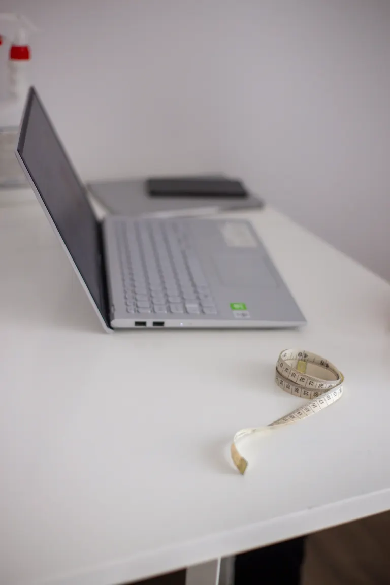 Laptop aberto em uma mesa branca ao lado de uma fita métrica enrolada.