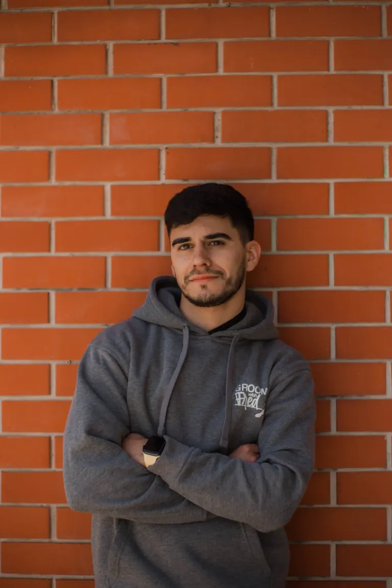 Homem jovem usando moletom cinza com capuz, braços cruzados, apoiado em parede de tijolos vermelhos, olhando para a câmera.