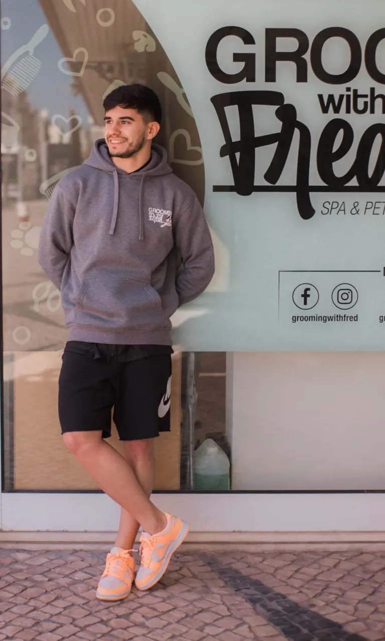 Homem jovem de camisa cinza com capuz e bermuda preta, usando tênis bege com detalhes em cinza, posando com uma perna...