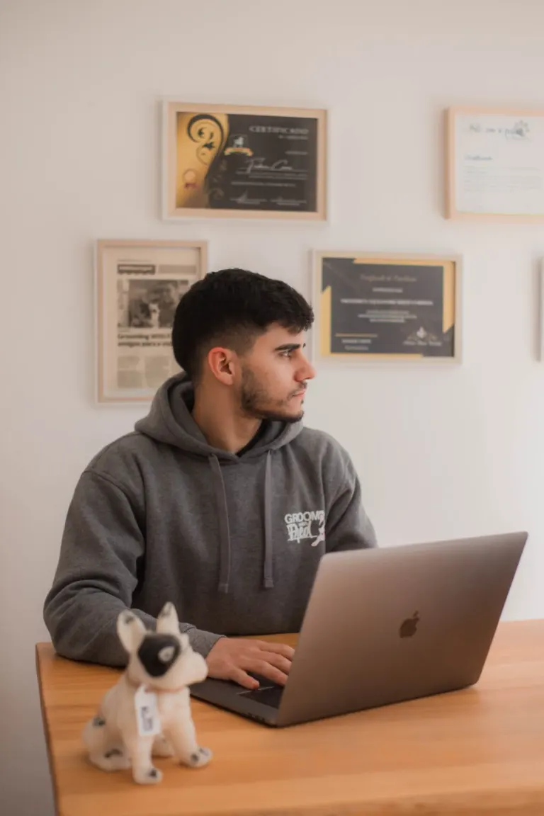 Homem sentado em uma mesa de madeira usando um laptop MacBook, com um brinquedo de cachorro na frente, atrás uma parede...