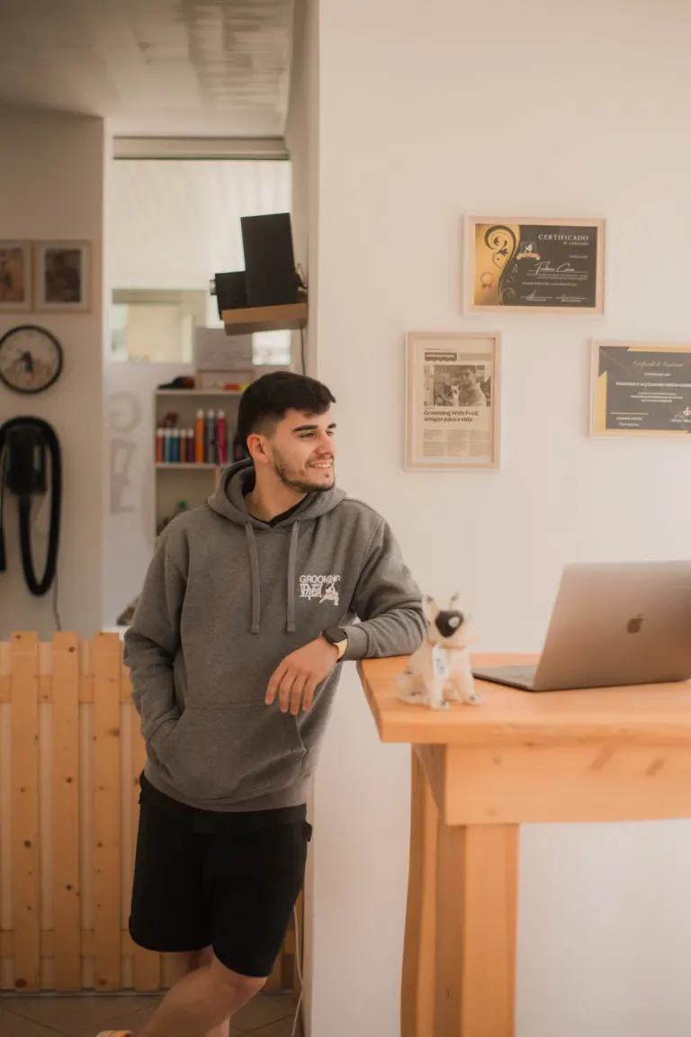 Jovem sorridente com camiseta de moletom cinza com logo de pet shop, usando relógio e bermuda preta, apoiado em balcão de...