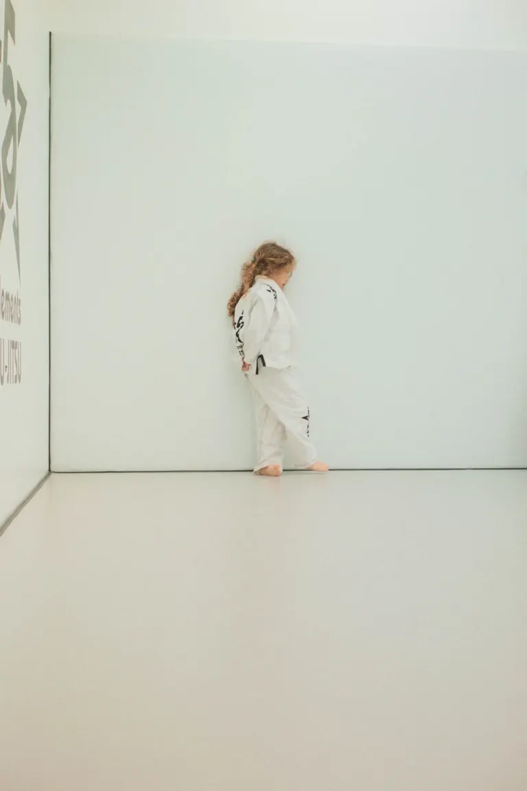 Menina de kimono branco com detalhes pretos, de cabelos castanhos, sentada contra uma parede branca, olhando para baixo,...