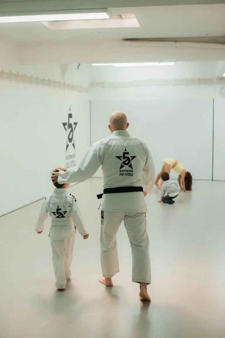 Instrutor de Jiu-Jitsu ensina uma criança na aula, enquanto duas outras pessoas praticam técnicas no chão em um dojo de...