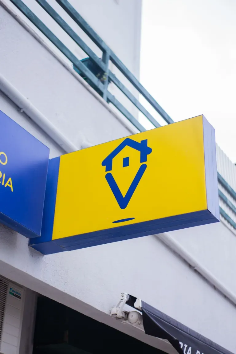 Placa de sinalização com fundo amarelo e desenho de uma casa estilizada no centro, com estrutura azul ao redor, fixada na...