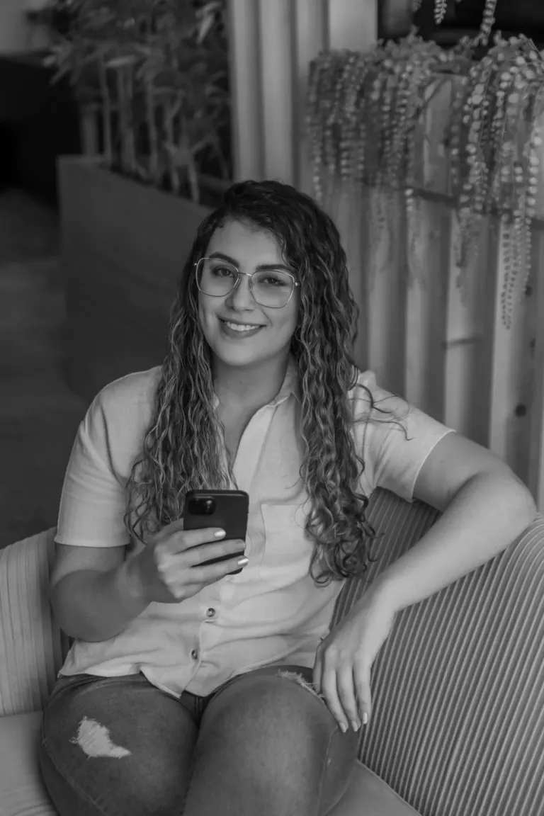 Mulher com cabelos cacheados, usando óculos, sorrindo enquanto segura um telefone móvel, sentada em um sofá no interior...
