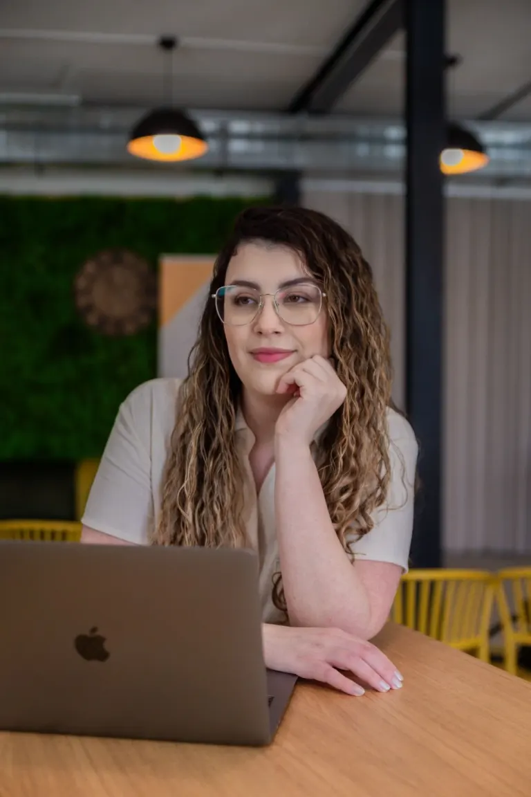 Mulher com cabelos cacheados e óculos, sentada em frente a um laptop, com expressão pensativa, no interior de um espaço...