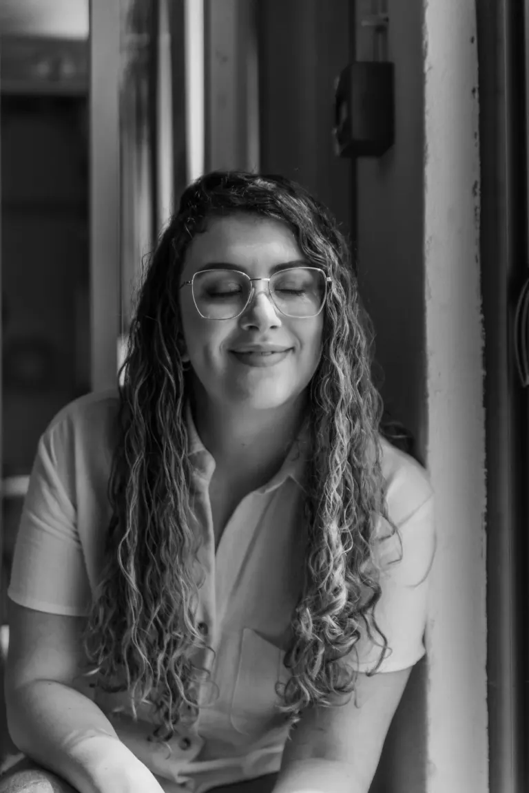 Retrato em preto e branco de uma mulher com cabelo cacheado, óculos e expressão serena, próxima a uma janela em um...