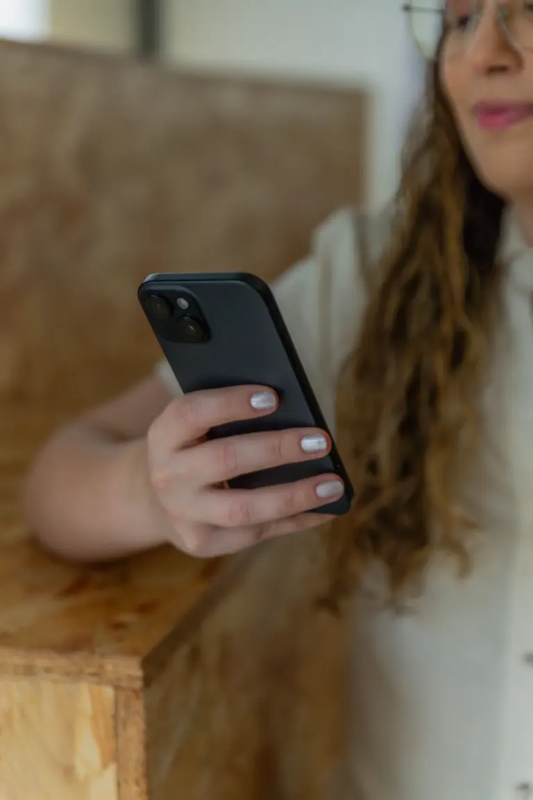 Pessoa segurando um smartphone preto com tela visível, ao fundo uma mulher com cabelo cacheado, óculos e blusa branca,...