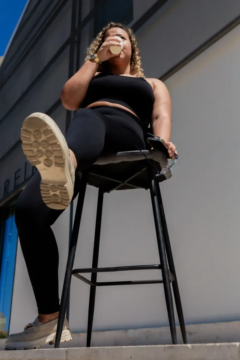 Mulher sentada em uma cadeira alta de costas, usando roupas pretas, tomando uma bebida e segurando óculos, vista de baixo...