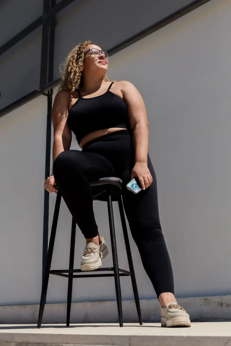 Mulher com cabelo cacheado, óculos escuros, vestindo blusa preta e calça preta, sentada em uma cadeira alta ao ar livre,...