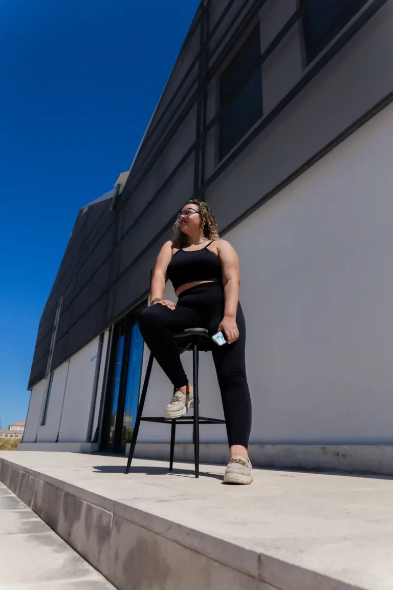 Mulher de sorriso descontraído usando roupa preta, sentada em banquinho na calçada de frente para uma parede moderna, sob...