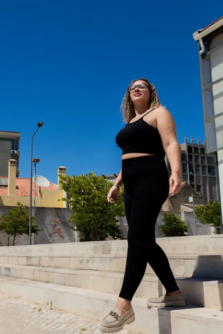 Mulher de cabelo cacheado, usando óculos de sol, manequim preto, calça preta, tênis bege, caminhando ao ar livre sob céu...