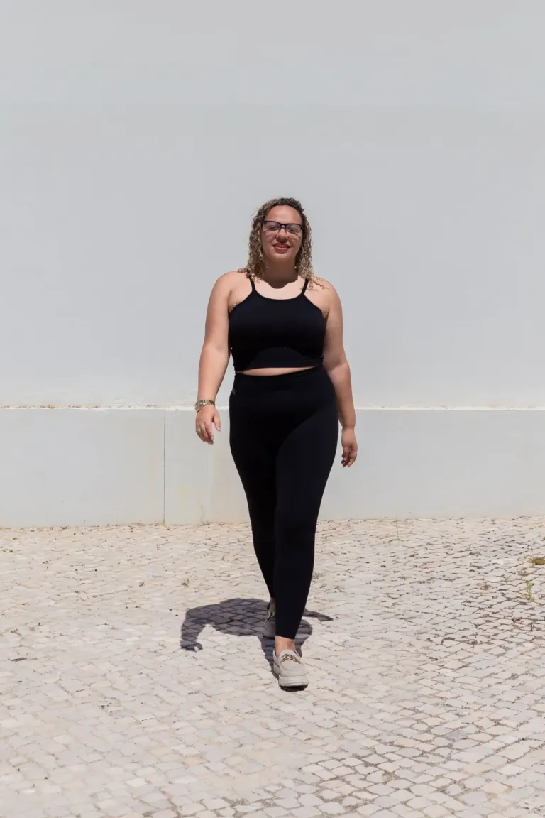 mulher de cabelo cacheado usando roupas de ginástica preta caminhando em calçada de pedra, com fundo branco