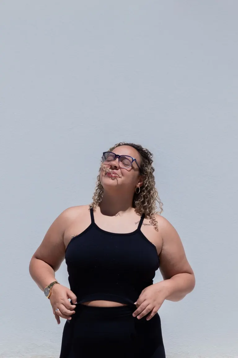 Mulher com cabelos cacheados longos, usando óculos de grau, camiseta preta e calça preta, posando com os olhos fechados...