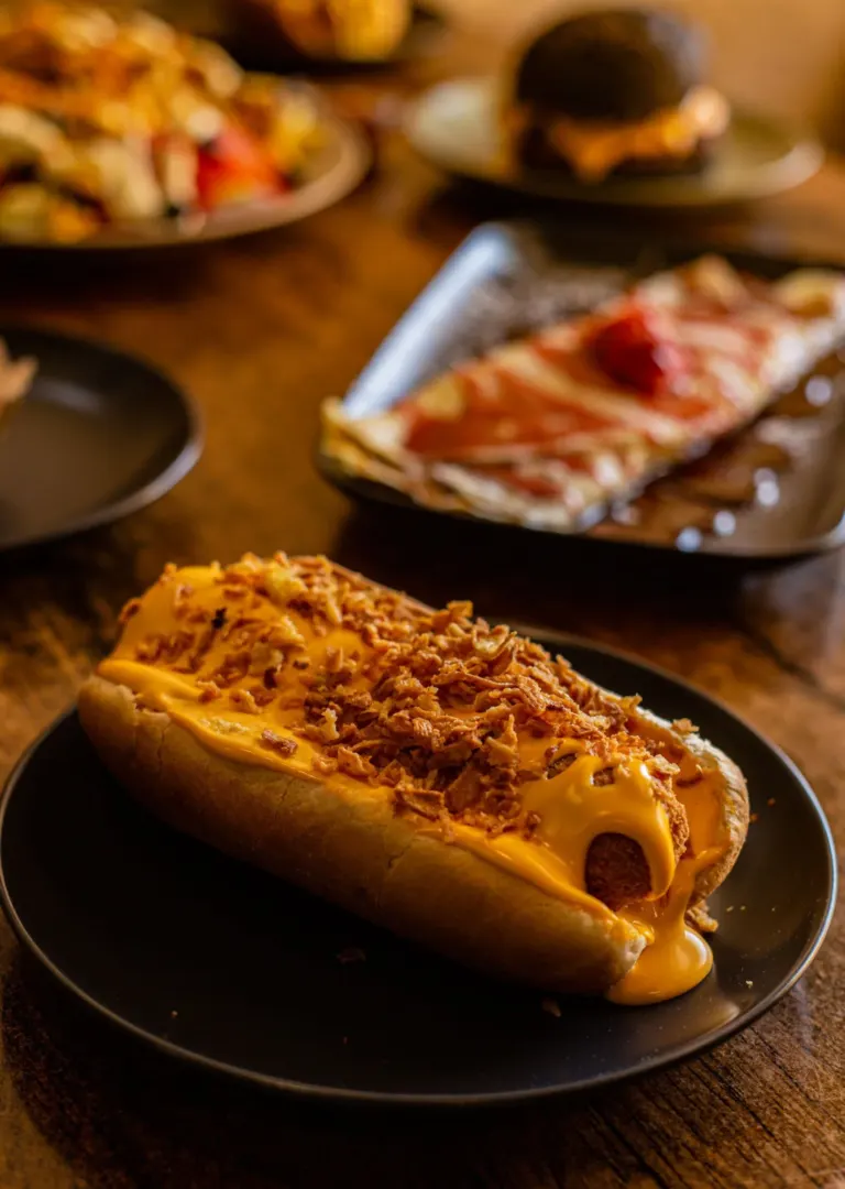 Cachorro-quente com queijo e bacon em um prato preto, ao fundo uma pizza e outras comidas em pratos pretos
