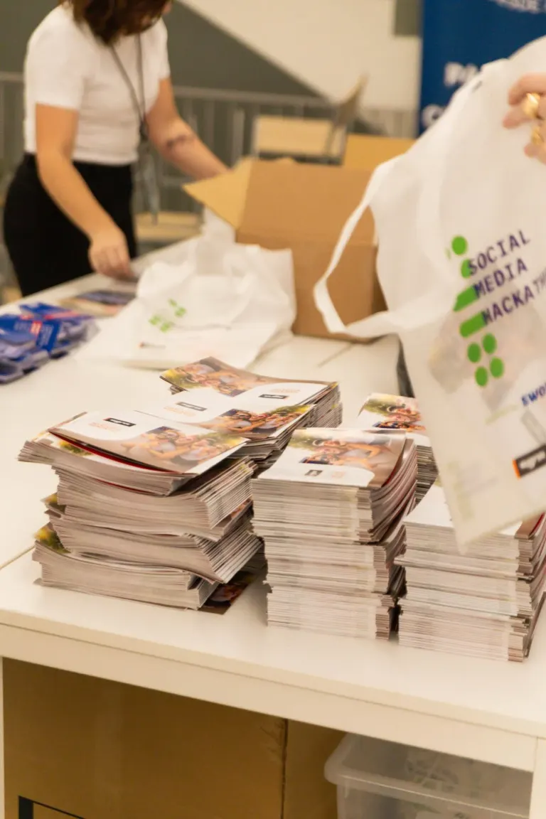 Pilhas de panfletos sobre uma mesa, com uma pessoa ao fundo abrindo uma caixa. Disparos de eventos de divulgação ou...
