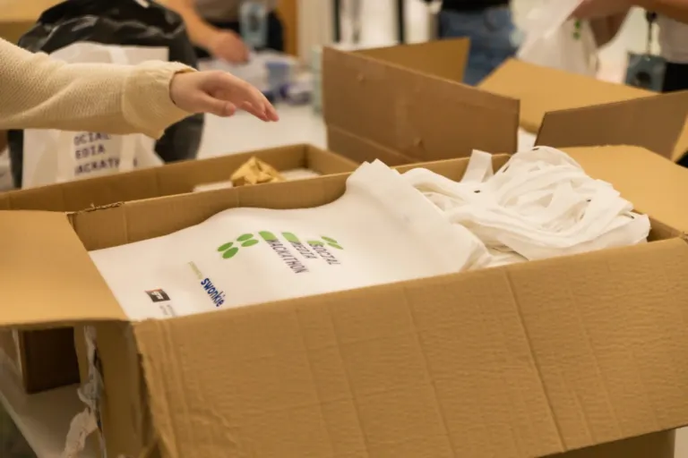 Caixa de papelão com sacolas brancas e fita de papel, pessoas ao fundo embalando objetos.