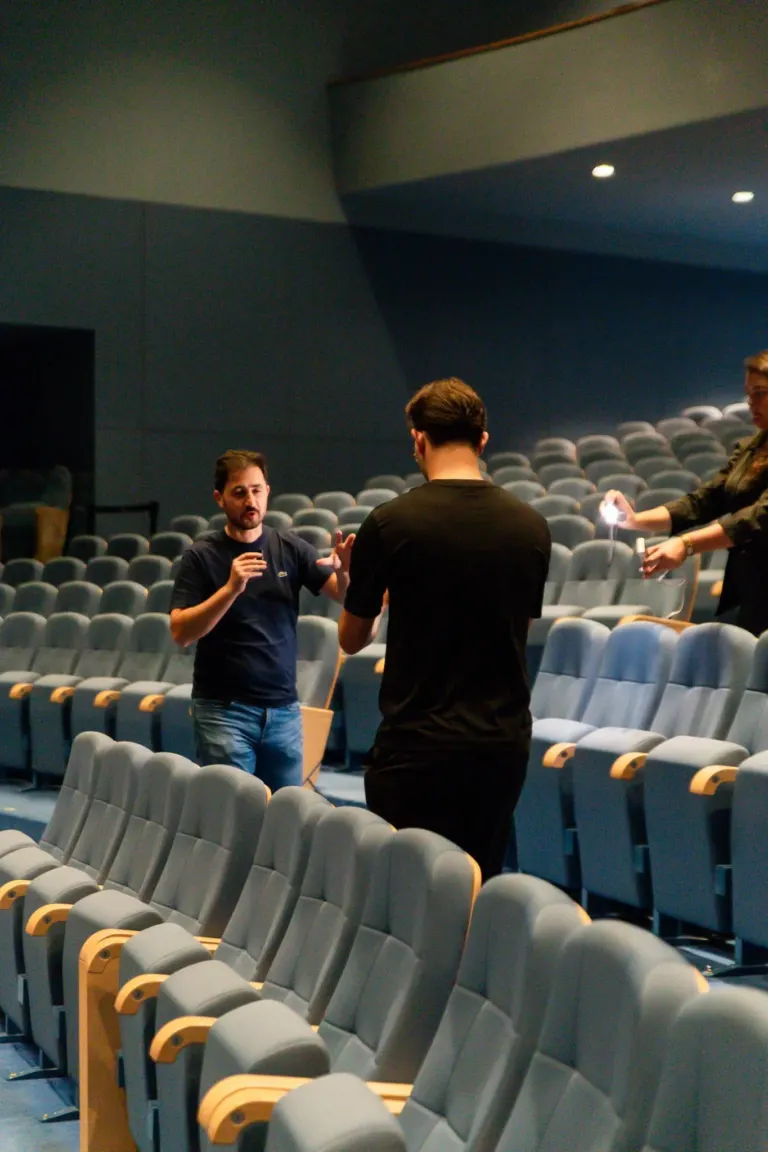 Homem discutindo com outro homem em uma sala de cinema vazia, enquanto uma mulher ao lado segura uma lanterna.