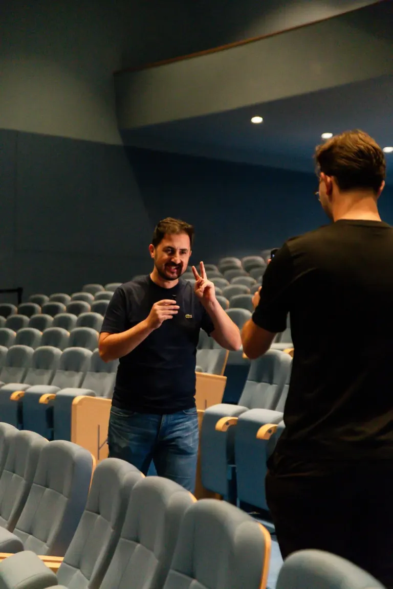 Duas pessoas em um teatro vazio, uma delas fazendo um sinal de paz enquanto sorri para a câmera e a outra pessoa tirando...