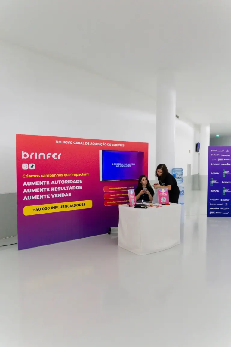 Estande de uma campanha de marketing digital com duas pessoas, uma sentada e outra de pé, usando celulares, próximo a uma...