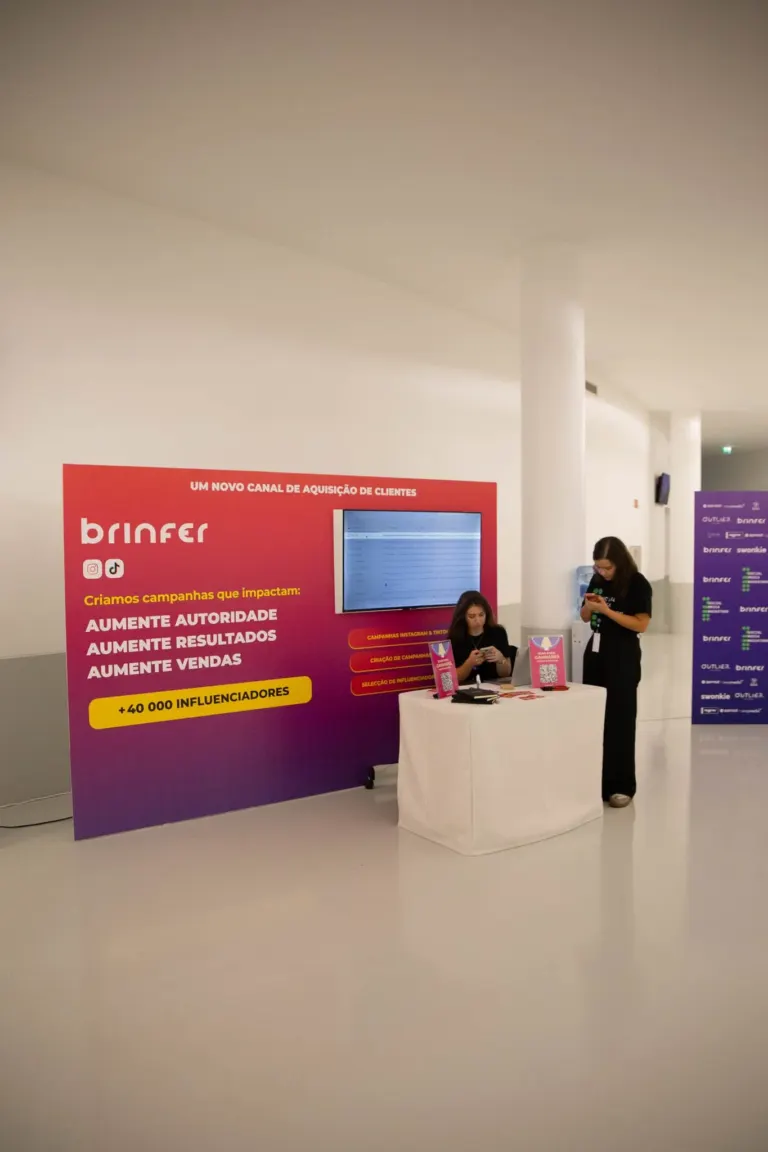 Stand de divulgação da Brinser com uma parede vermelha, uma mesa com material de divulgação e duas mulheres usando...
