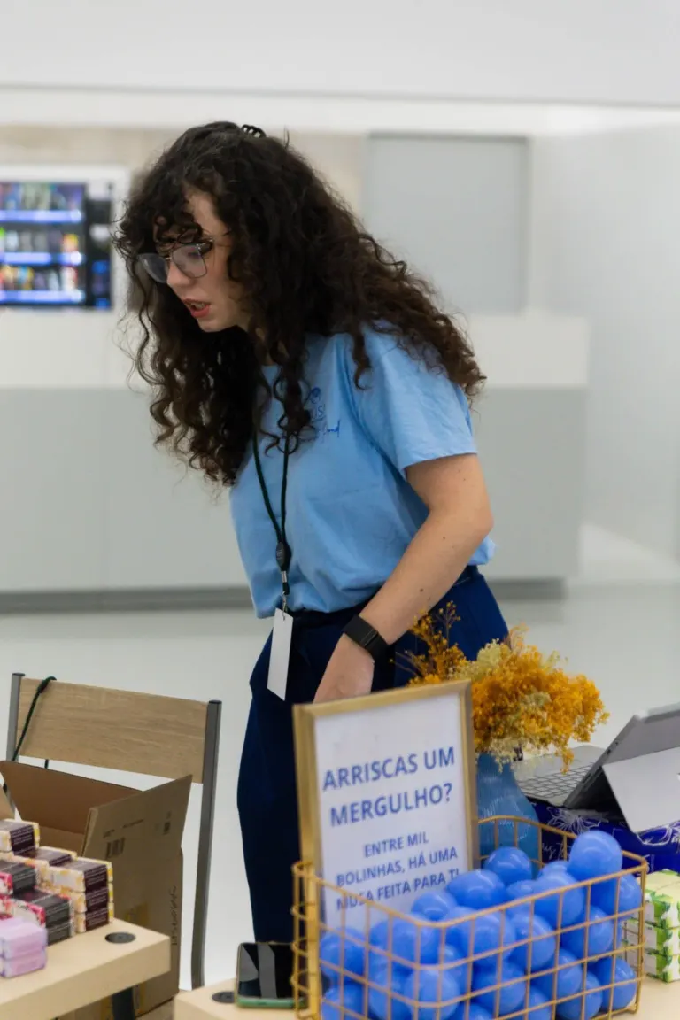 Mulher com cabelo cacheado e óculos, usando camiseta azul, em uma loja ou feira, ao lado de uma mesa com produtos e um...