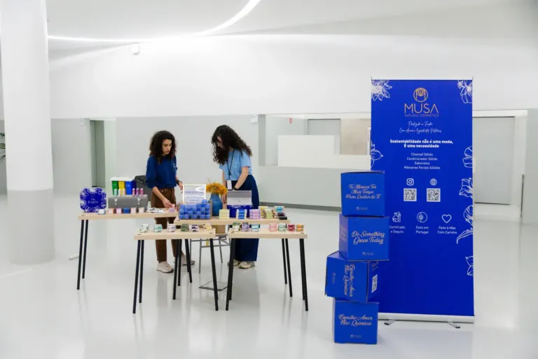 Duas mulheres em uma mesa de exibição de produtos de cosméticos naturais, com caixas azuis e coloridas, dentro de um...