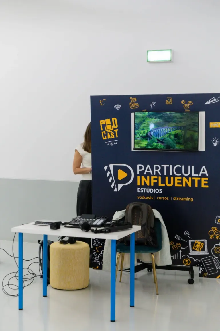 Estande de divulgação de um podcast de estudos, com banner preto e amarelo, uma televisão mostrando uma paisagem verde,...