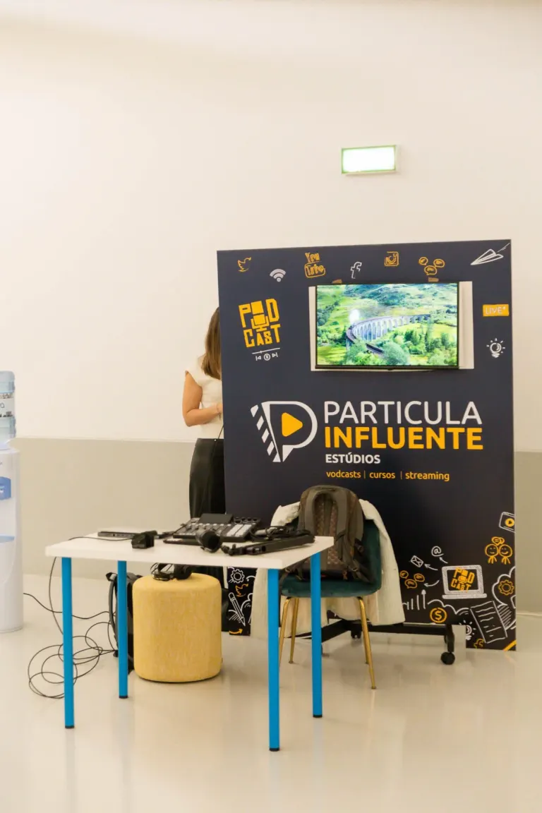 Estande de divulgação de uma escola de podcasts chamada 'Partícula Influente', com um painel colorido, televisão exibindo...